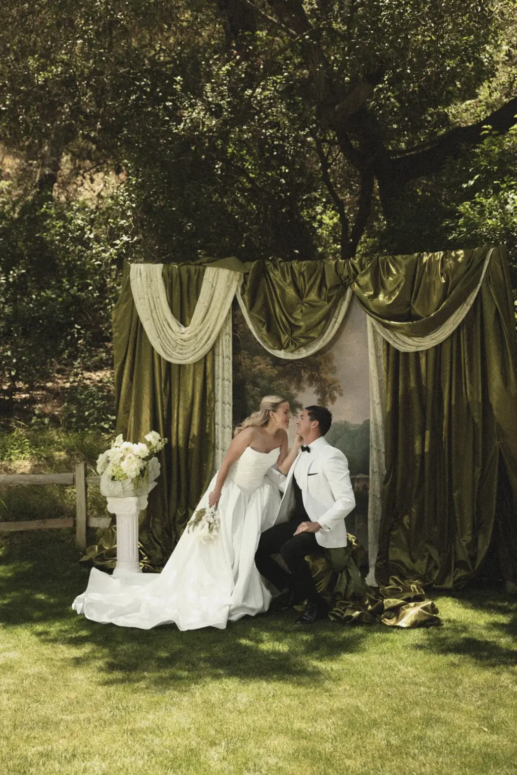 Bride and groom lean toward one another in front of an olive silk draped altar with a painted landscape backdrop at an outdoor garden ceremony.