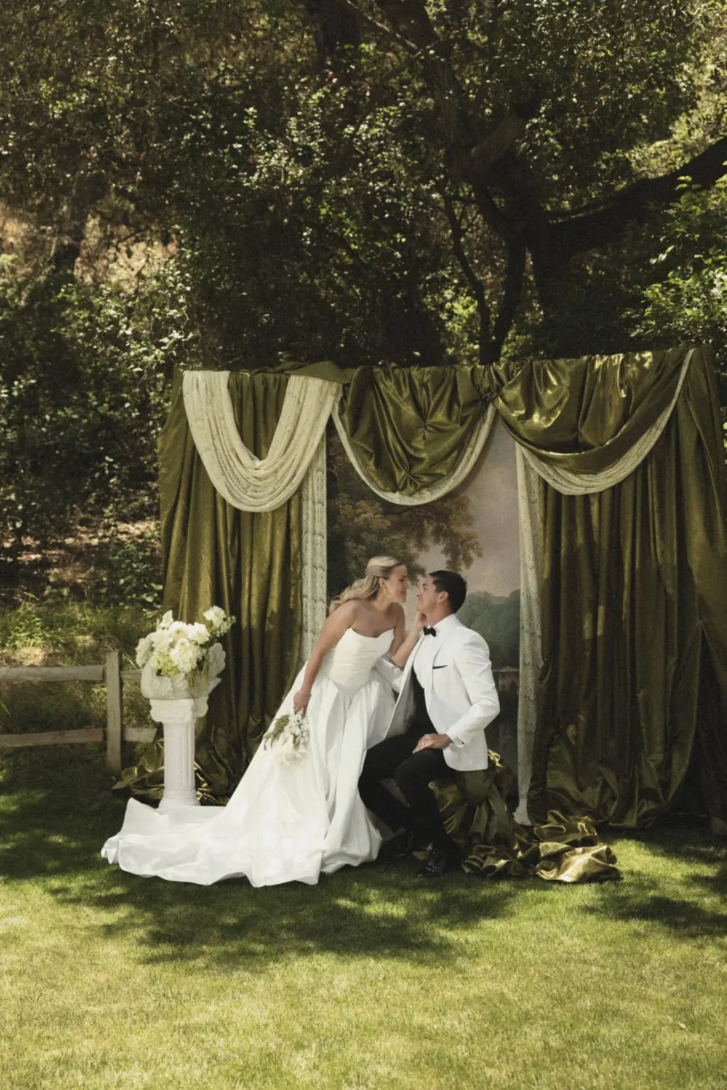 Bride and groom kiss before a draped green velvet altar backdrop on a garden lawn