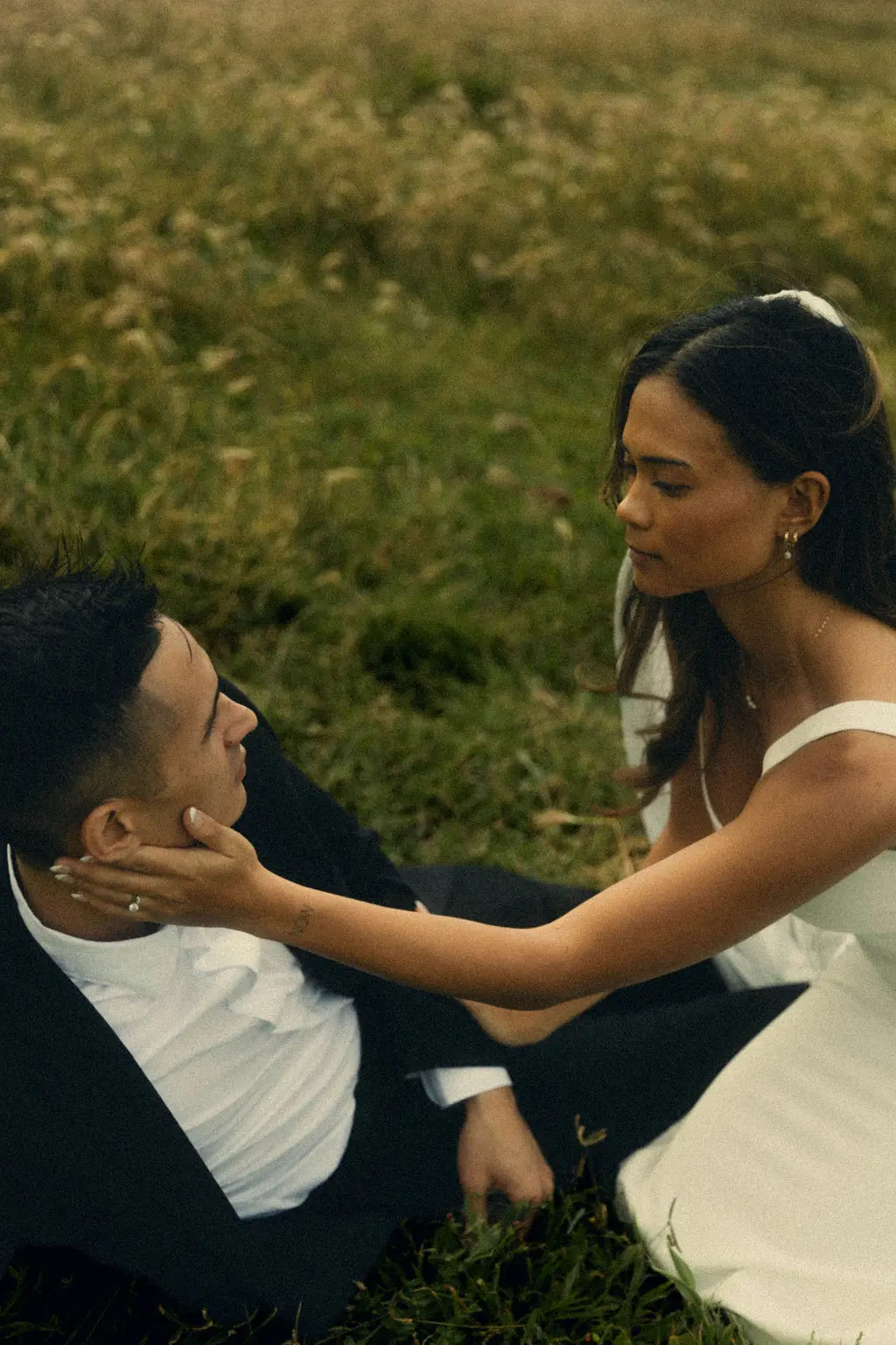 Bride cradles the groom's face as they sit together in a sunlit meadow of tall grass.
