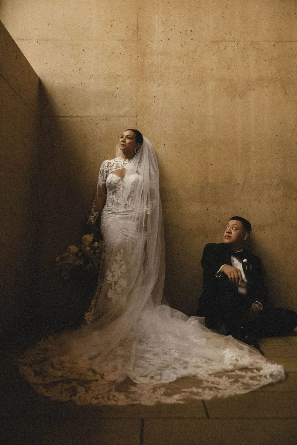 Bride in a long-sleeved lace gown and cathedral veil stands against a warm concrete wall while the groom sits on the floor beside her train.