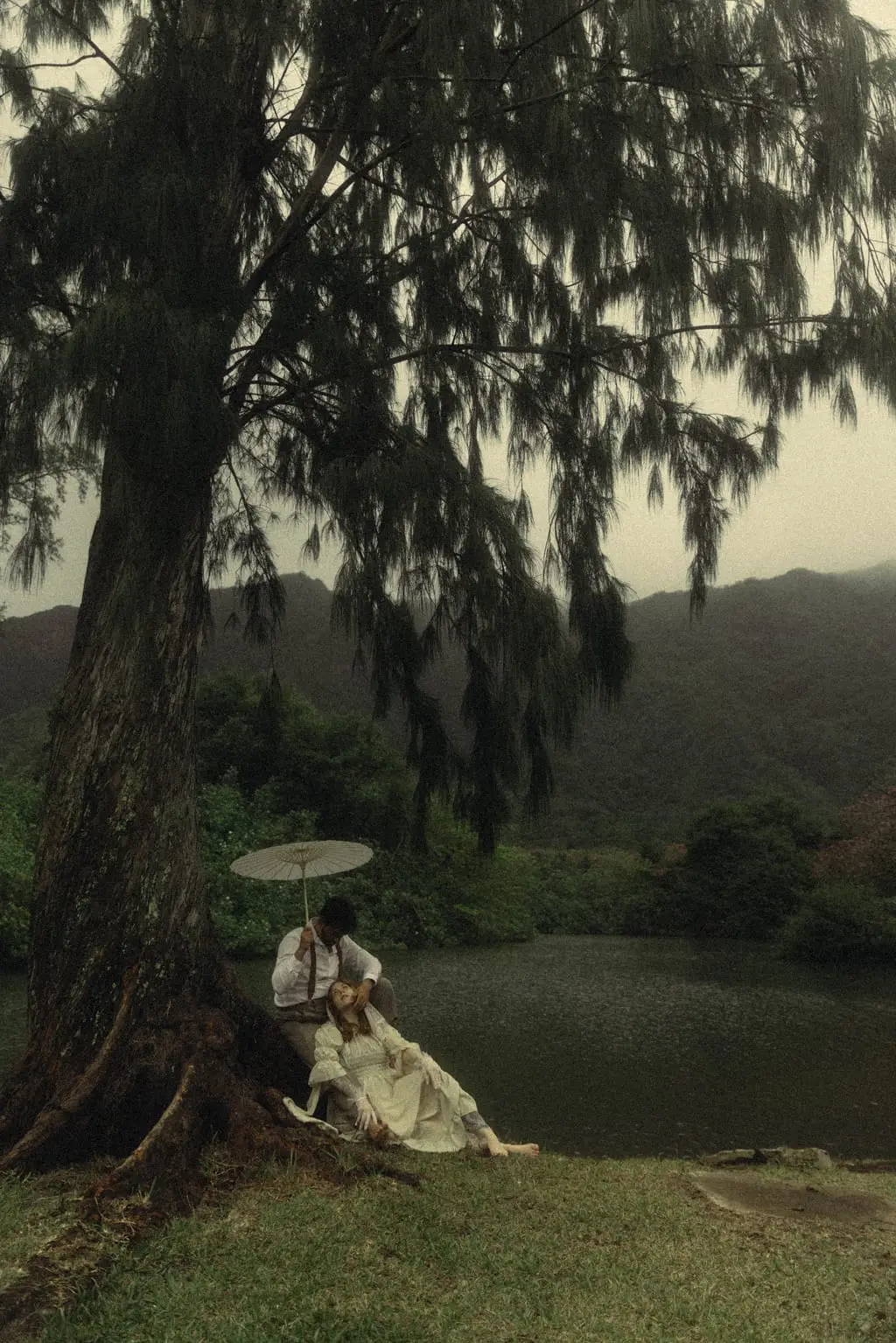 Couple rest together beneath a windswept ironwood tree with a paper parasol beside a mountain pond during a misty Hawaii elopement.