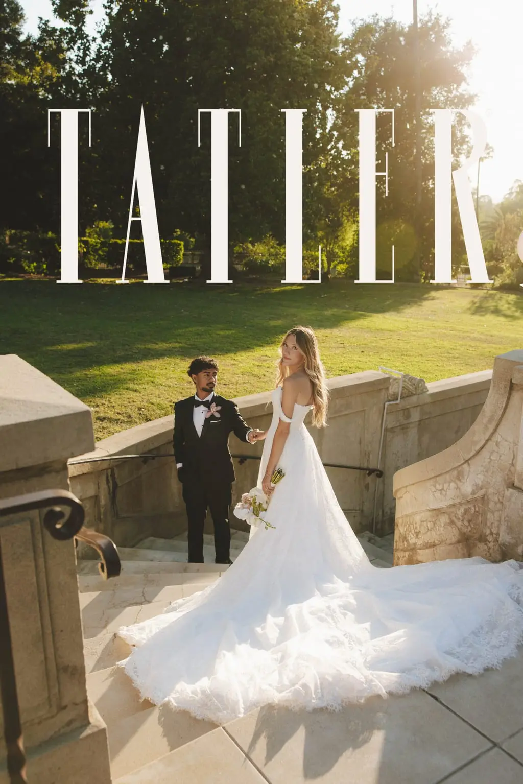 Bride in a lace ball gown with a long cathedral train looks back over her shoulder at her groom on the stone steps of a sunlit garden estate, featured in Tatler.