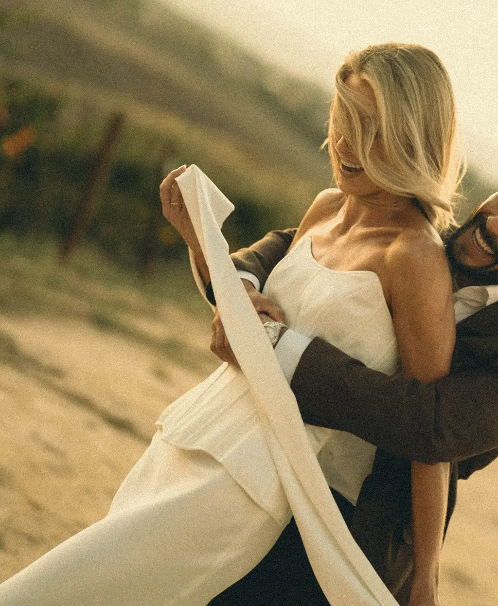 Groom lifts laughing bride against golden hillside light at a warm outdoor wedding.
