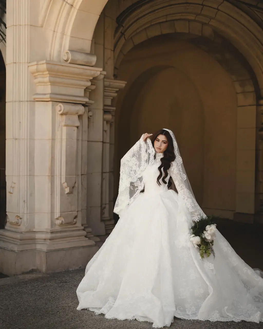 Bride in a lace ballgown and veil lifts her veil beneath a grand stone archway and columns